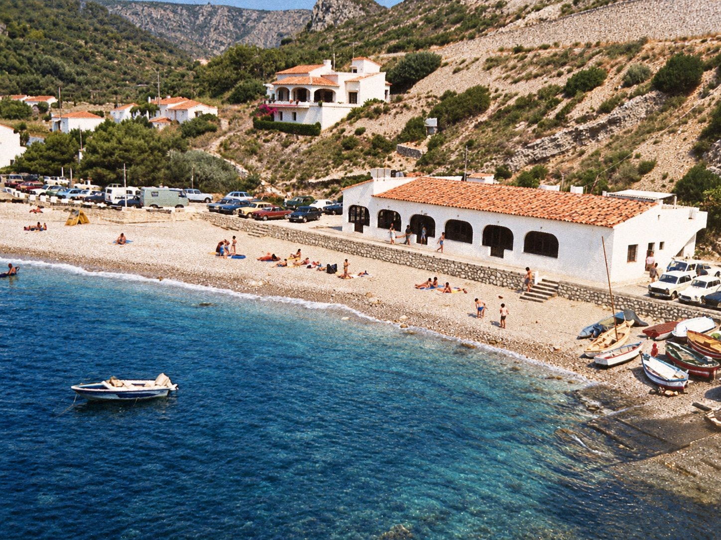 Old photograph: the beach with bathers, beached boats and a white arched building with a tiled roof.